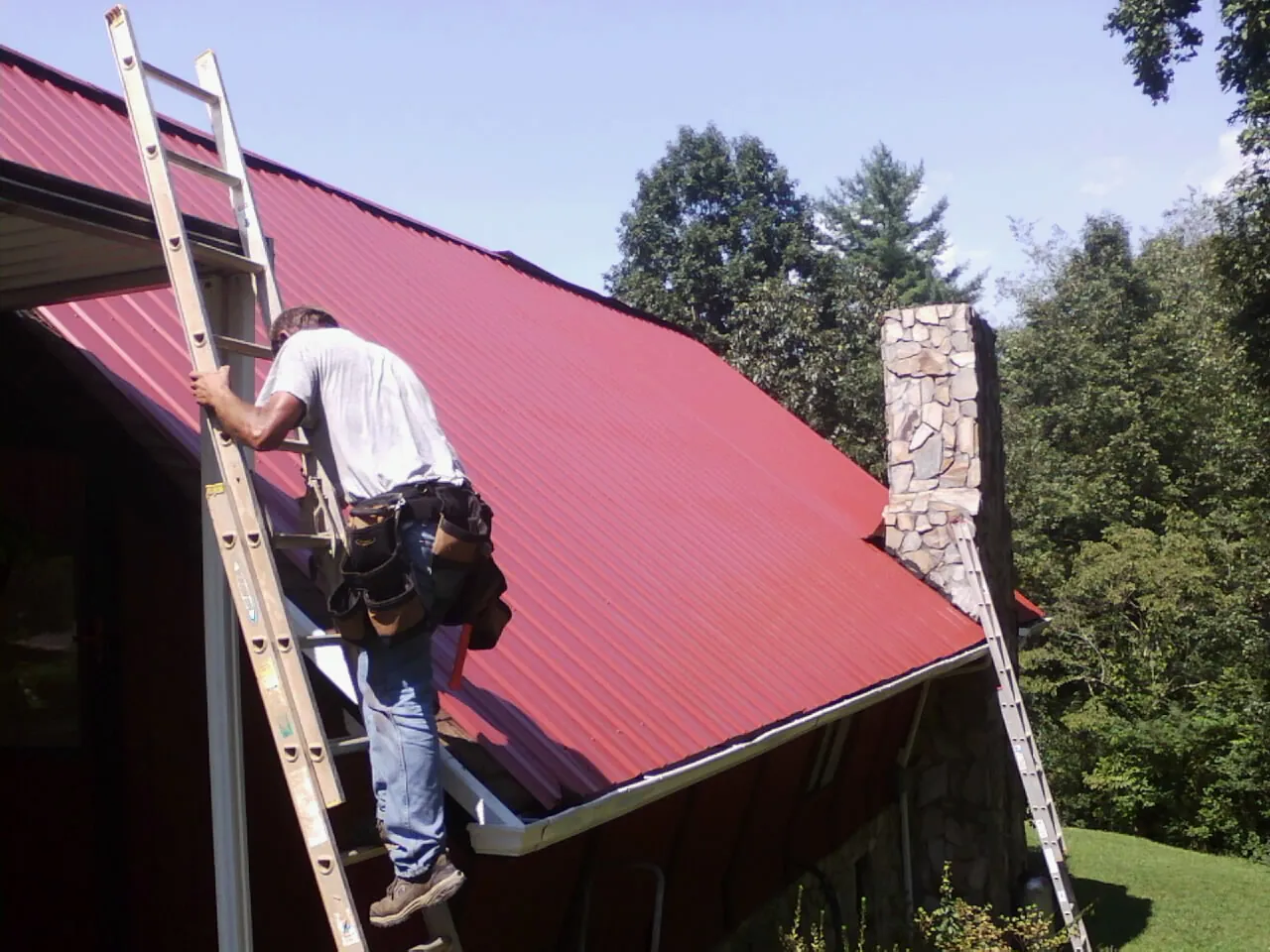 Roofer on a ladder beside a red metal roof and stone chimney — Soffit & Fascia Repair by Roofing Brothers in Bryce